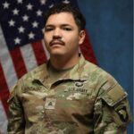A military serviceman posing in uniform with the American flag in the background.