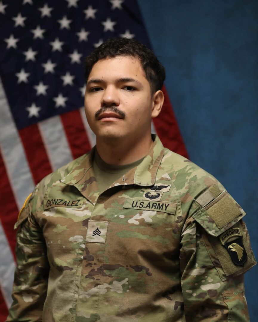 A military serviceman posing in uniform with the American flag in the background.